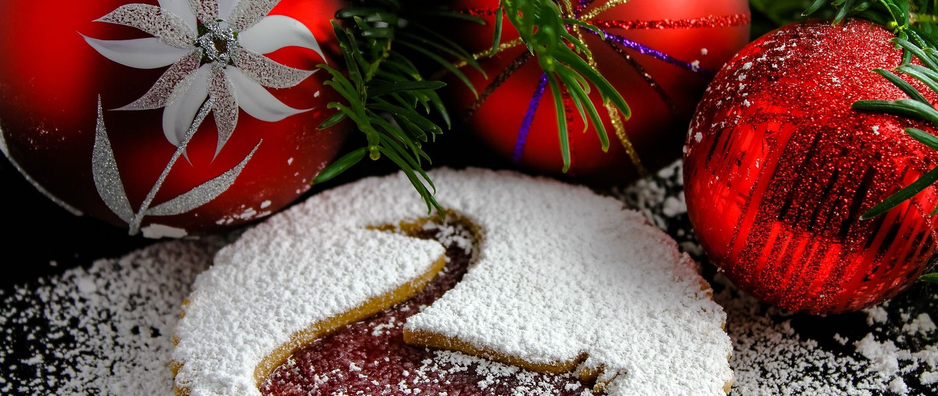 Weihnachtliche Szene mit roten Christbaumkugeln und einem mit Puderzucker bestäubten Plätzchen.