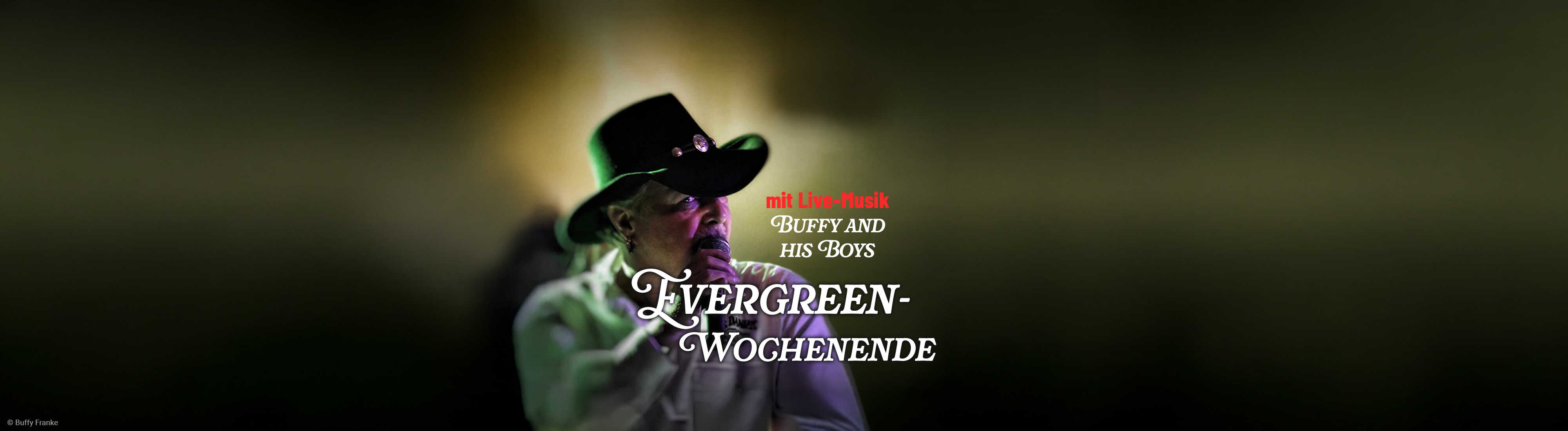 Ein Musiker mit schwarzem Cowboyhut singt ins Mikrofon und sorgt vor dunkler Bühnenkulisse für stimmungsvolle Live-Musik-Atmosphäre.
