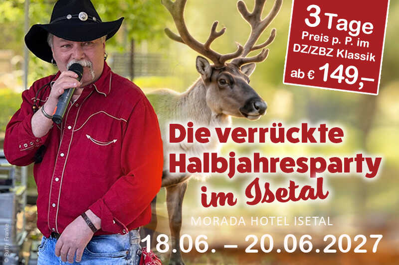 Ein Sänger im roten Westernhemd und schwarzen Cowboyhut steht mit Mikrofon neben einem Rentier in sommerlicher Naturkulisse.
