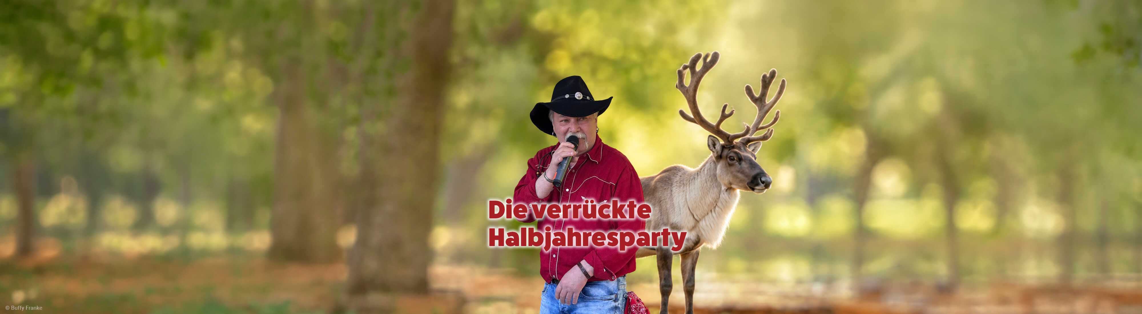 Ein Sänger im roten Westernhemd und schwarzen Cowboyhut steht mit Mikrofon neben einem Rentier in sommerlicher Naturkulisse.

