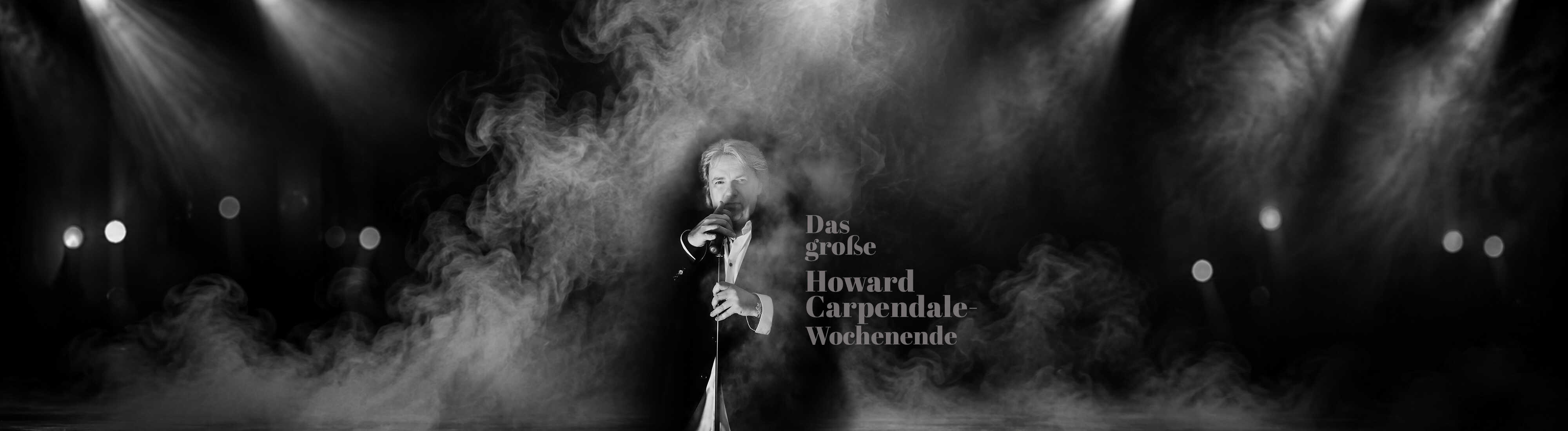 Ein Sänger im eleganten Anzug steht mit Mikrofon in einer schwarz-weißen, neblig inszenierten Bühnenkulisse und vermittelt klassische Konzertatmosphäre.
