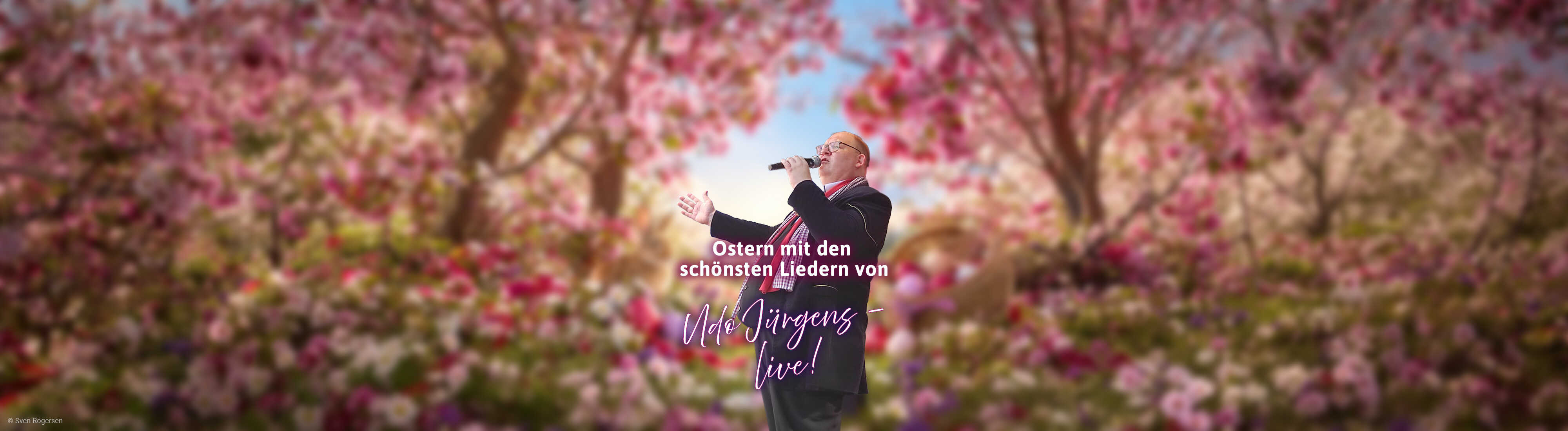 Ein Sänger mit Mikrofon steht in frühlingshafter Osterkulisse mit blühenden Zweigen, Osterhasen und farbenfroher Dekoration.
