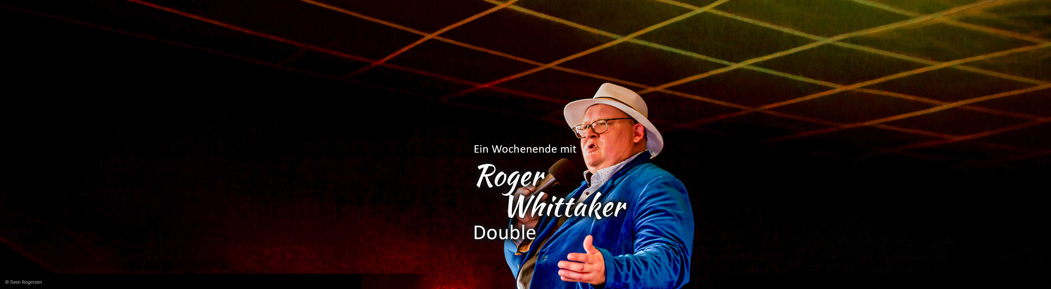 Ein Sänger im blauen Sakko und weißen Hut steht mit Mikrofon auf der Bühne vor einem dunklen, stimmungsvoll beleuchteten Hintergrund.