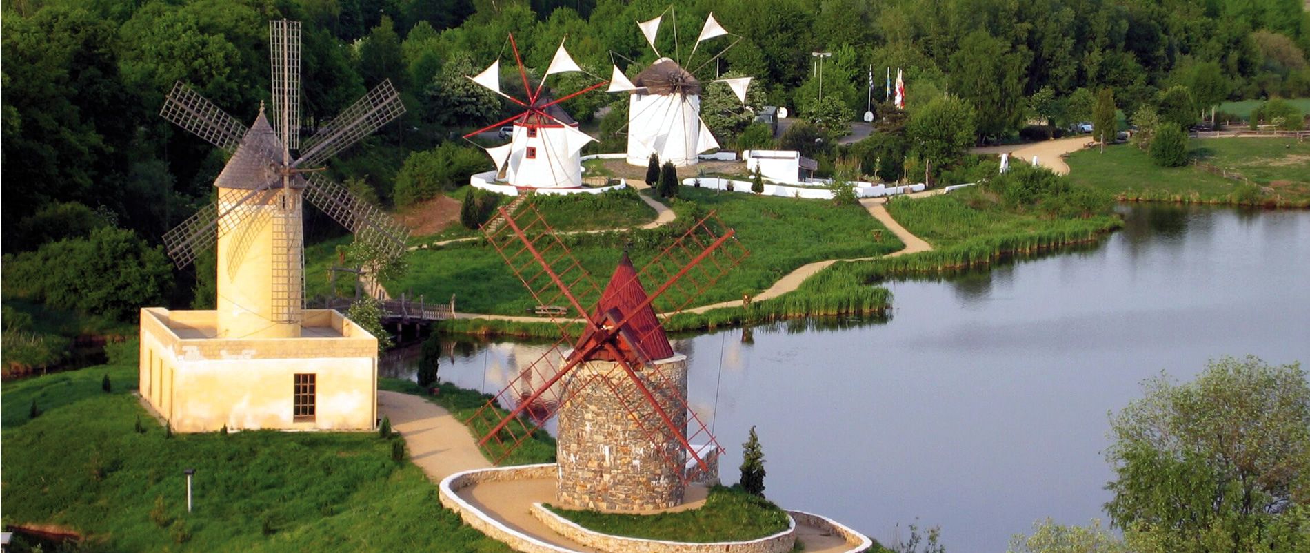 Panorama von drei traditionellen Windmühlen an einem Seeufer, umgeben von üppigem Grün.