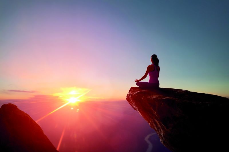 Person meditiert im Lotussitz auf einem Felsen mit atemberaubendem Sonnenuntergang im Hintergrund.