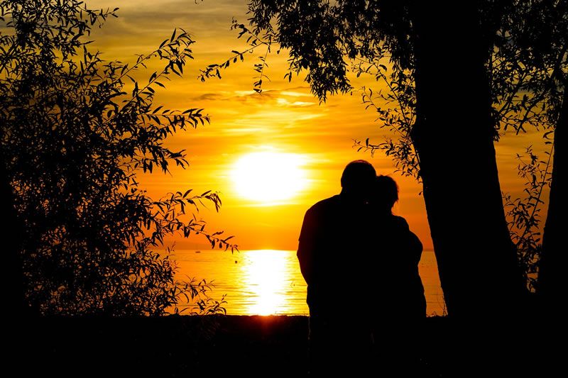 Silhouette eines Paares vor einem Sonnenuntergang am See, umgeben von Bäumen.