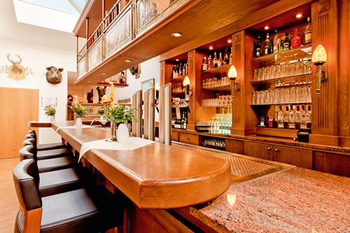 Innenansicht einer stilvollen Bar mit Holzmöbeln, einer langen Theke, Barhockern und Regalen mit Getränken und Gläsern.