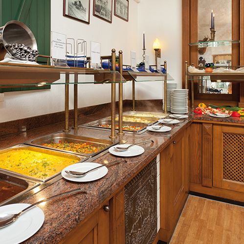 Buffet mit verschiedenen warmen Gerichten in Metallschalen, darunter Gemüse- und Fleischgerichte. Teller und Löffel liegen bereit.