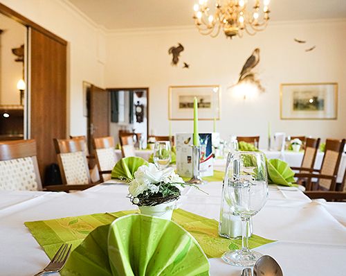 Festlich gedeckter Tisch in einem elegant eingerichteten Raum mit Kronleuchter und Wanddekoration, grüne Servietten und Weingläser auf den Tischen.