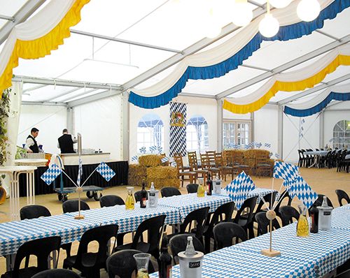 Oktoberfest-Zelt mit blau-weißen Tischdecken, dekoriert mit bayerischen Fahnen und Strohballen.