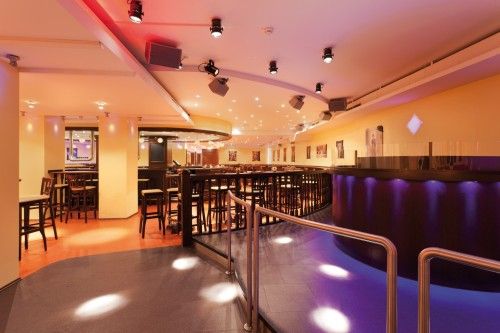 Innenansicht einer stilvoll beleuchteten Bar mit hohen Tischen und Stühlen, modernem Design und stimmungsvoller Beleuchtung.