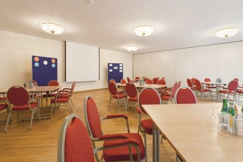 Konferenzraum mit roten Stühlen und Tischen, einem Projektorbildschirm und Pinnwänden.