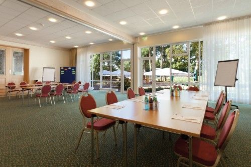 Heller Konferenzraum mit großen Fenstern, Tischen, roten Stühlen und Flipcharts.