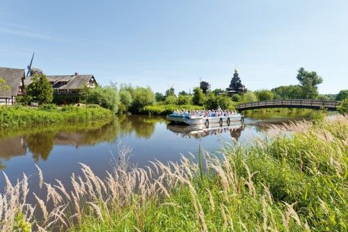 Grüne Landschaft mit einem Fluss, auf dem ein Touristenschiff fährt, umgeben von historischen Gebäuden und einer Brücke.