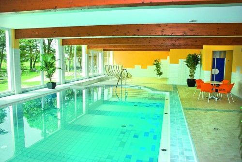 Innenansicht eines modernen Hallenbads mit großen Fenstern, einem Pool und orangefarbenen Stühlen.