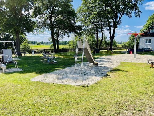 Spielplatz mit Rutsche, Schaukel und Wippe, umgeben von Bäumen und Wiese.