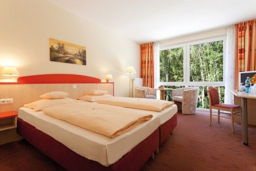 Helles Hotelzimmer mit Doppelbett, großem Fenster mit Blick ins Grüne, einem Schreibtisch und gemütlichem Sesselbereich.