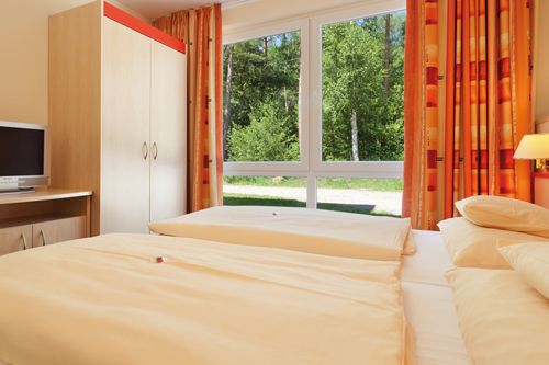 Helles Schlafzimmer mit zwei Einzelbetten, einem Schrank, einem Fernseher und großen Fenstern mit Blick auf einen Garten.