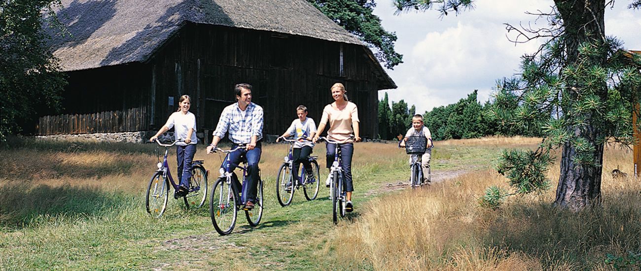 Eine Gruppe von Menschen fährt auf Fahrrädern einen ländlichen Weg entlang, neben einem Fachwerkhaus mit Strohdach.