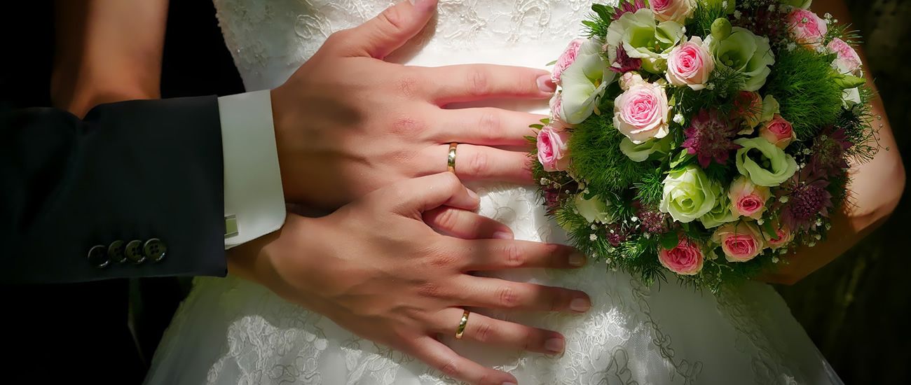 Verschränkte Hände eines Hochzeitspaars mit Eheringen auf dem Brautkleid, daneben ein Blumenstrauß.